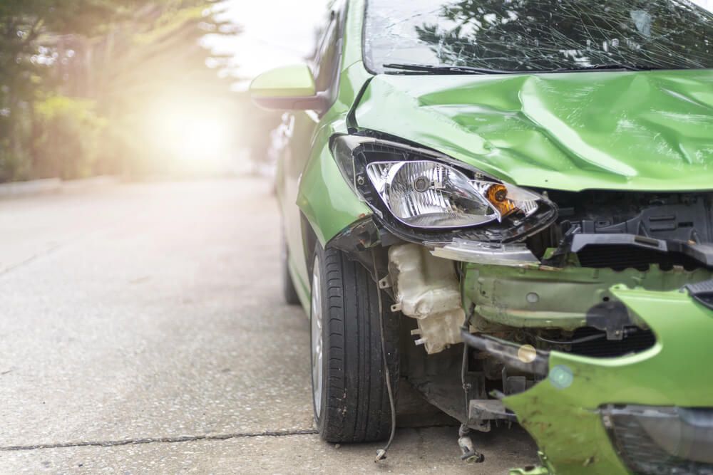 A green car with a damaged front end is parked on the side of the road.