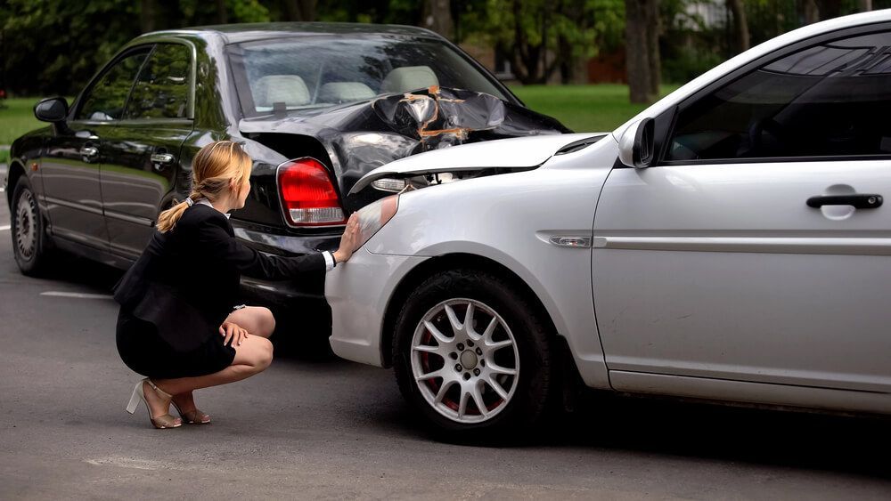 A person in a business suit crouches to inspect damage to the front bumper of a silver car that rear-ended a black car.