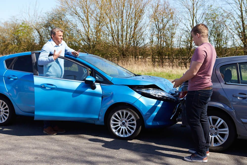 Two people stand by their damaged cars on a road, gesturing and discussing a collision during daylight.