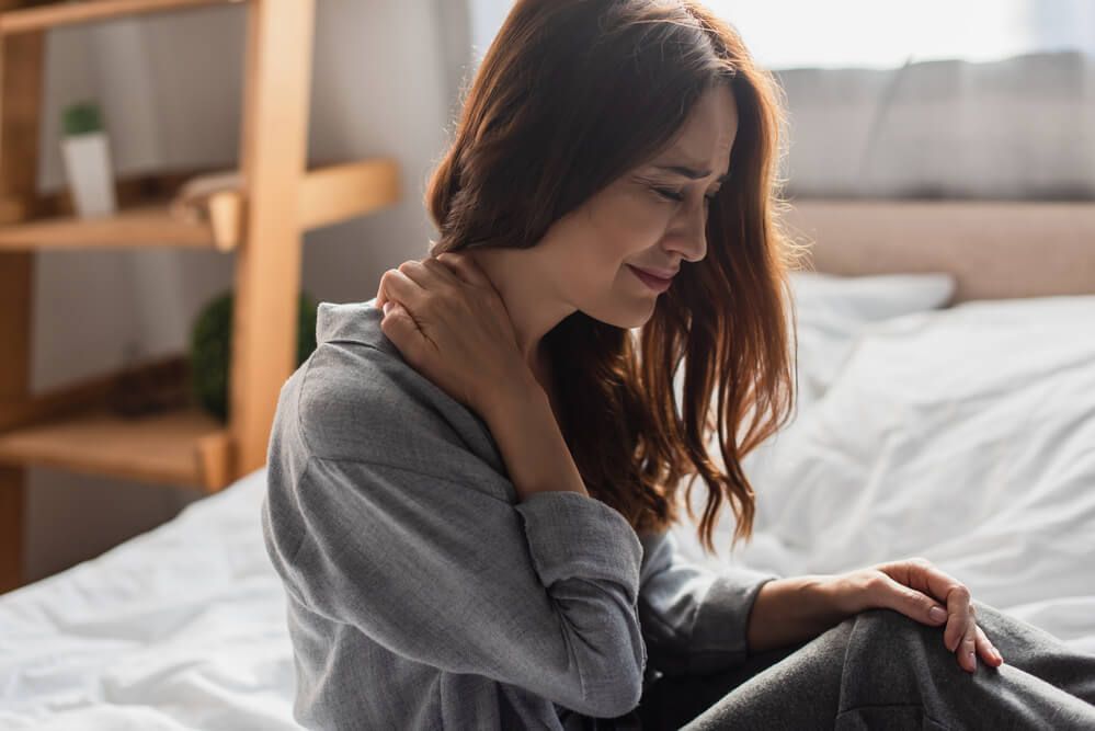 A person sits on a bed, clutching their neck and looking distressed while experiencing physical discomfort.