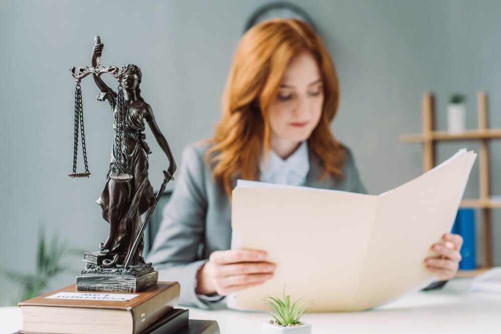 A woman is sitting at a desk with a statue of justice in the background.