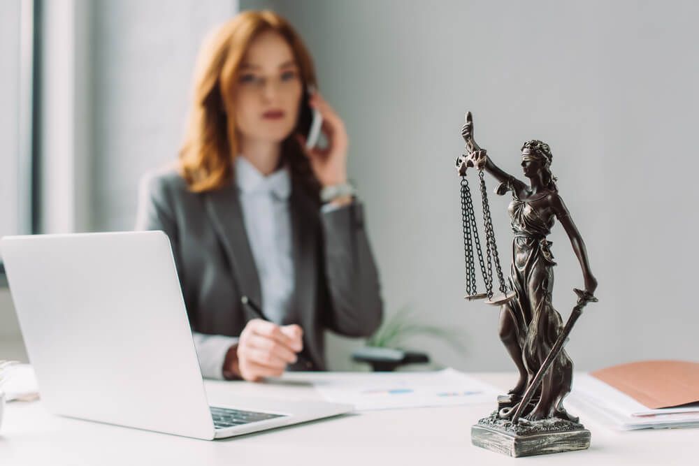 A woman is sitting at a desk talking on a cell phone next to a statue of justice.