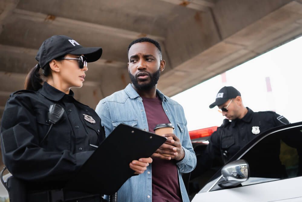 A police officer holding a clipboard talks to a man holding a coffee cup, with another officer nearby by a patrol car.