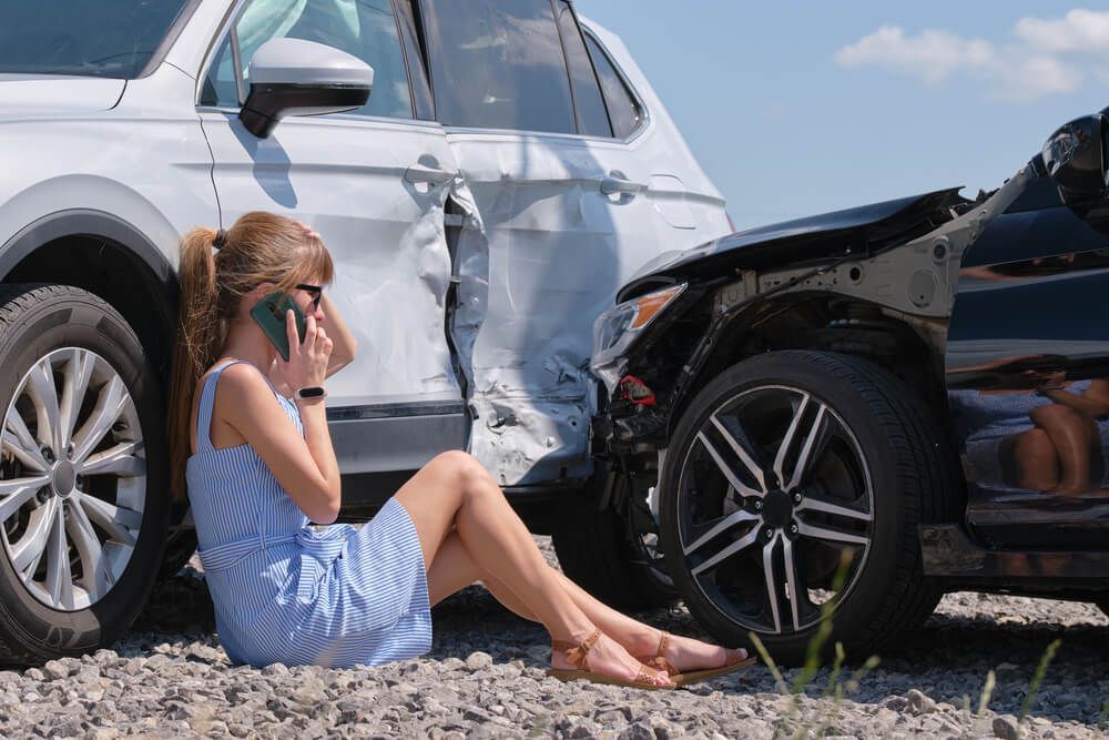 A person sitting on gravel against a damaged white car after a two-vehicle collision, using a cell phone.
