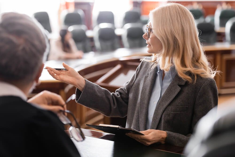 A woman is talking to a judge in a courtroom while holding a clipboard.