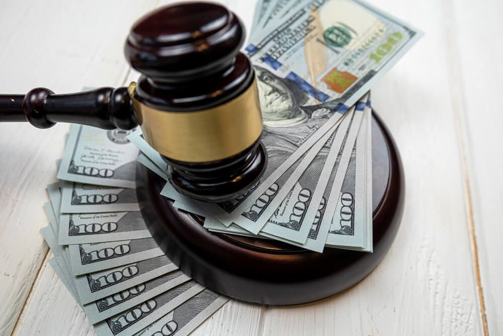 A wooden gavel rests on top of a stack of United States one-hundred-dollar bills on a white wooden surface.