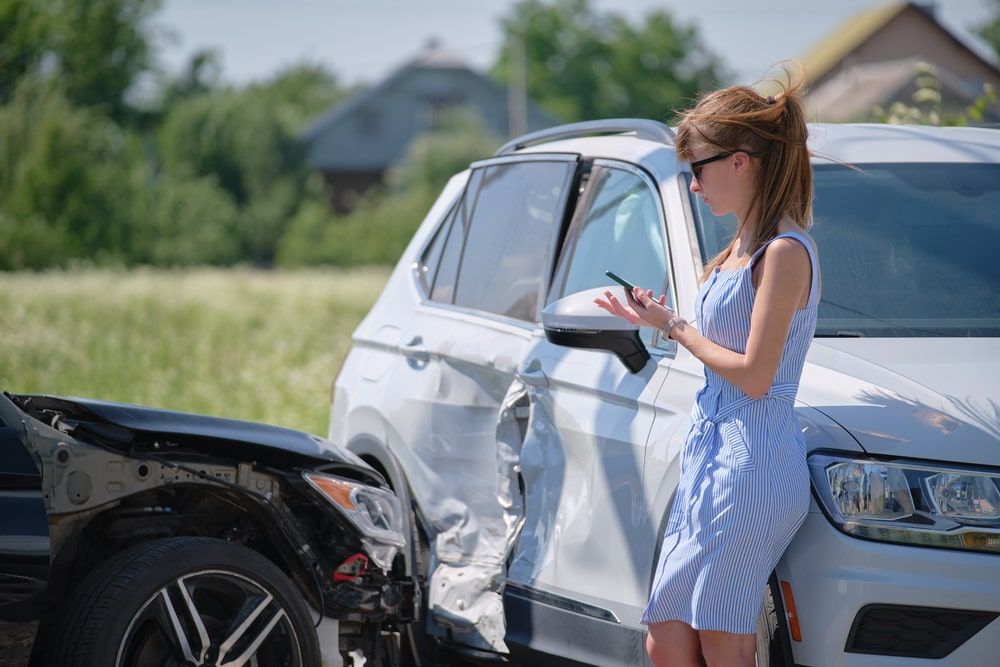 A woman is standing next to a damaged car and looking at her phone.