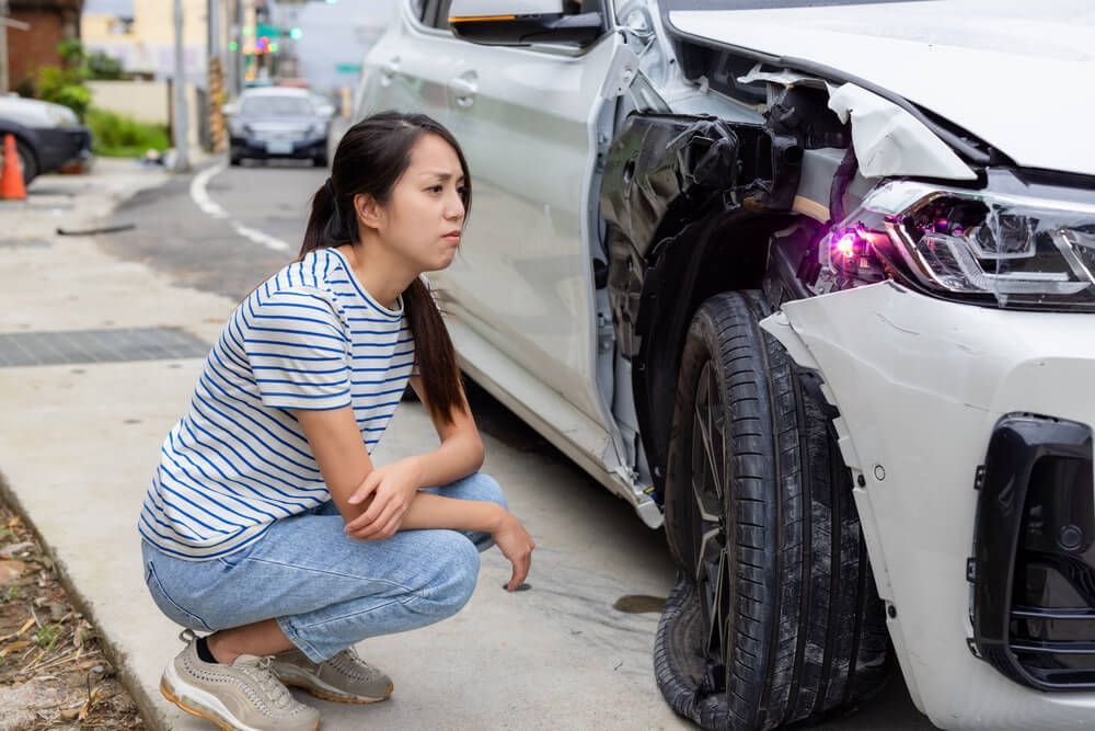 A woman is kneeling down next to a damaged car.