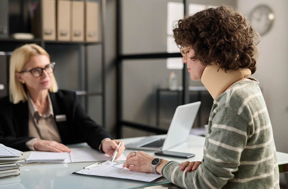 A professional in a blazer sits across from an individual wearing a neck brace at a desk to review documents.