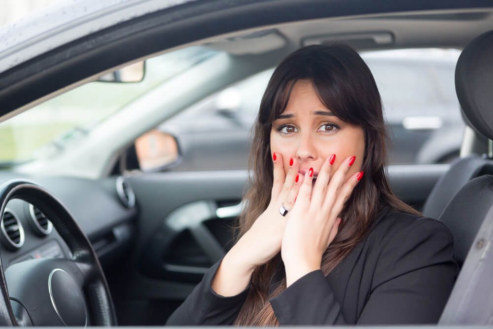 A person sits in the driver's seat of a car, hands pressed to their face with an expression of shock or surprise.