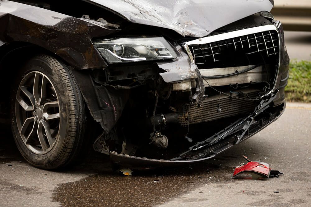 A black car with a broken bumper is parked on the side of the road.