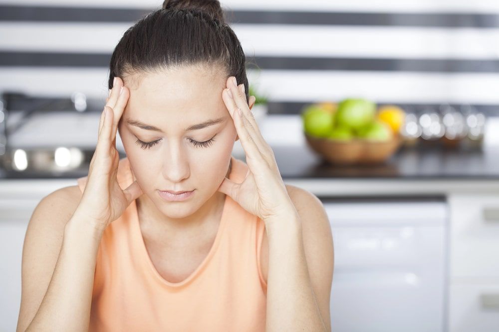 A woman is sitting in a kitchen with her hands on her head.