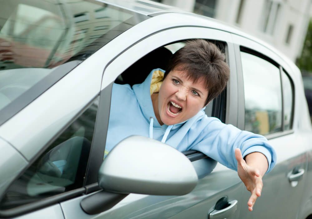 A person in a light blue hoodie shouting from the driver's seat of a car with an arm outstretched in frustration.