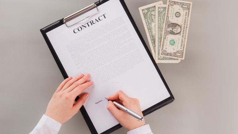 A person is signing a contract on a clipboard next to money.
