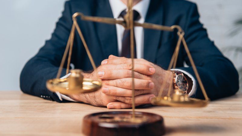 A man in a suit and tie is sitting at a table with his hands folded in front of a scale of justice.