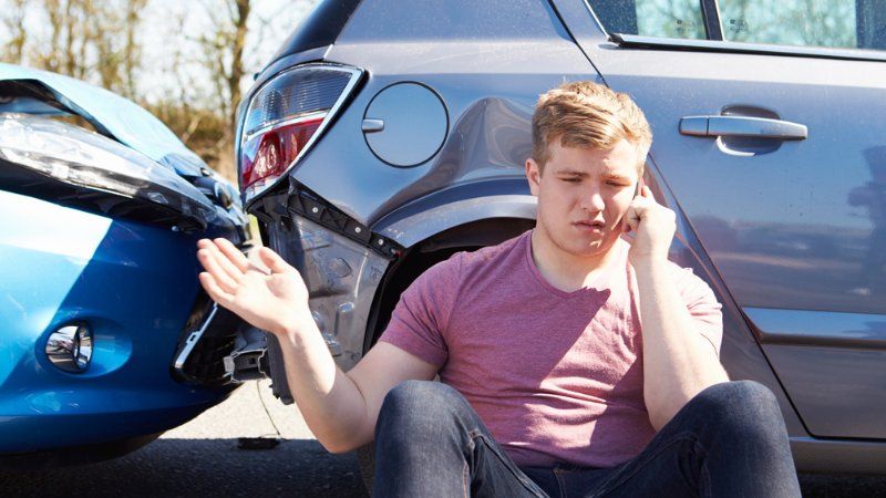 A man is sitting on the ground next to a damaged car talking on a cell phone.