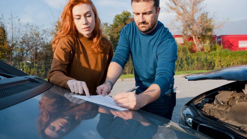 A man and a woman are looking at a piece of paper next to a broken down car.