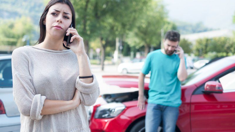 A woman is talking on a cell phone in front of a man and a red car.
