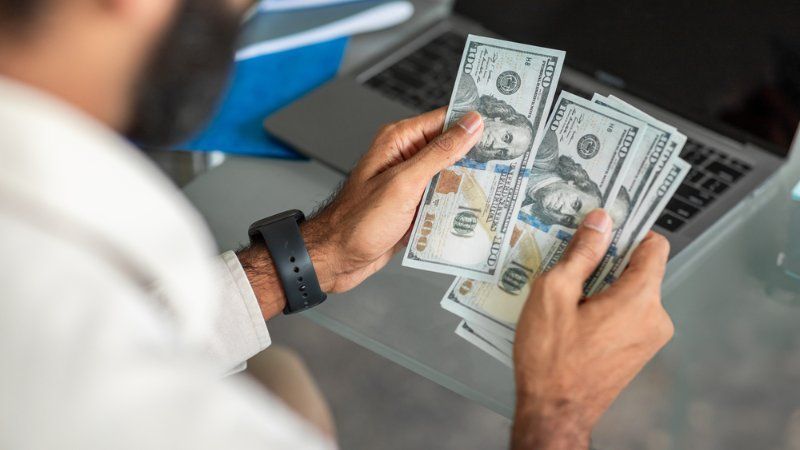 A person counts a stack of hundred-dollar bills while sitting at a laptop.