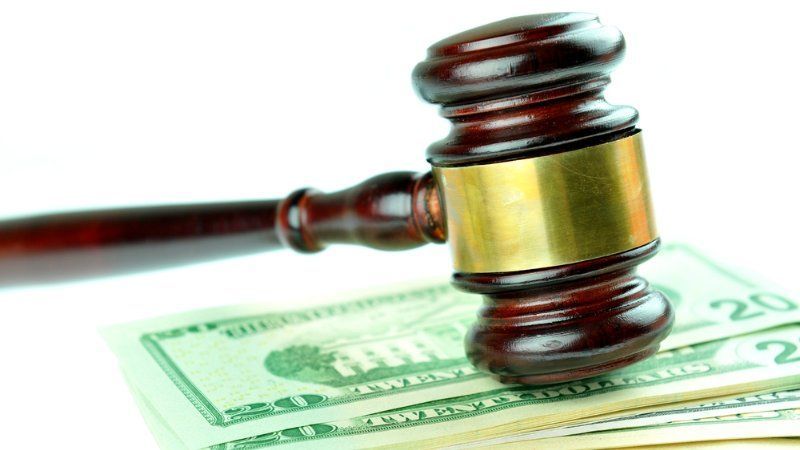 A wooden judge’s gavel resting on a stack of twenty-dollar bills against a white background.