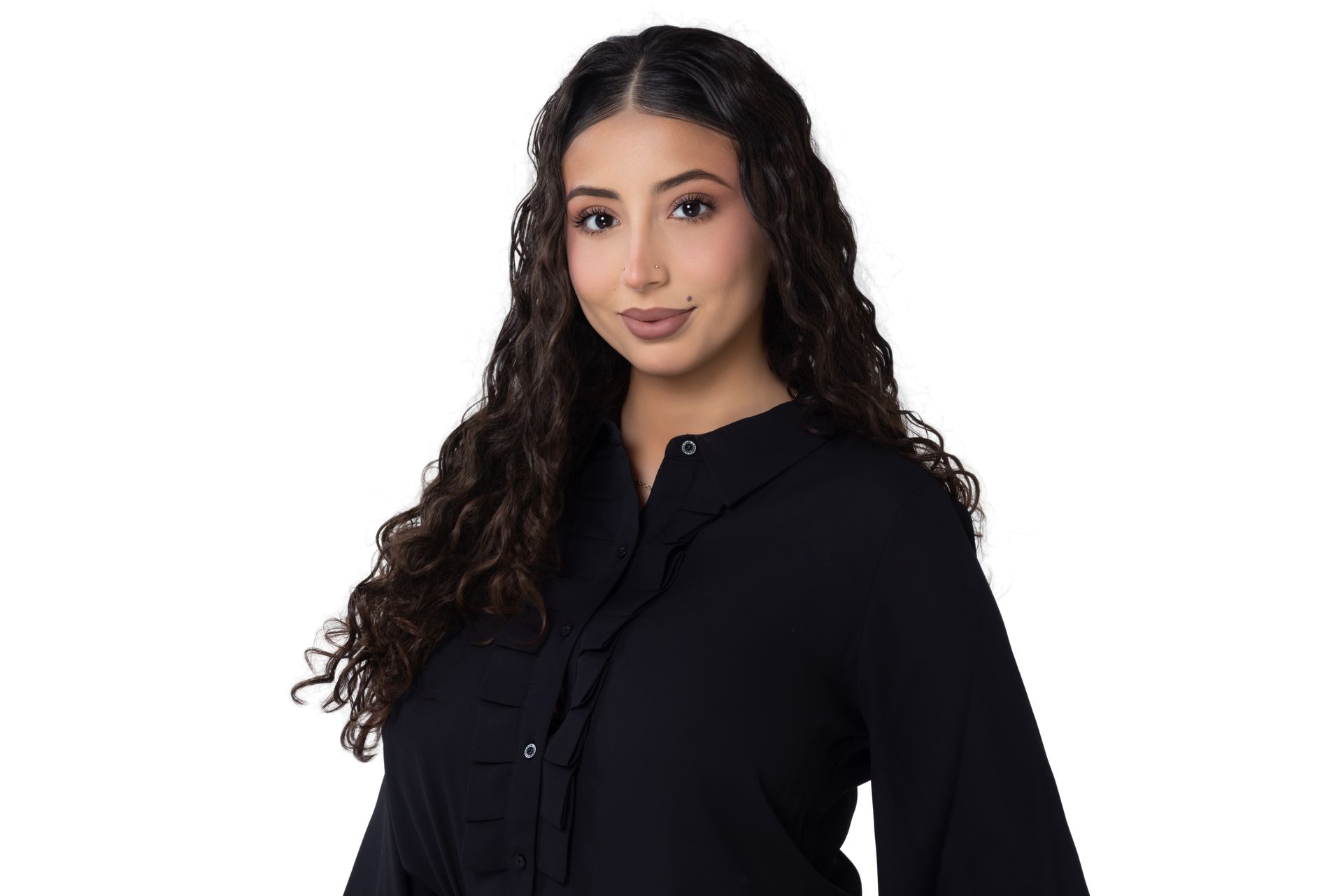 A person with long, curly dark hair, wearing a black button-down blouse with ruffled trim, posing against a white background.