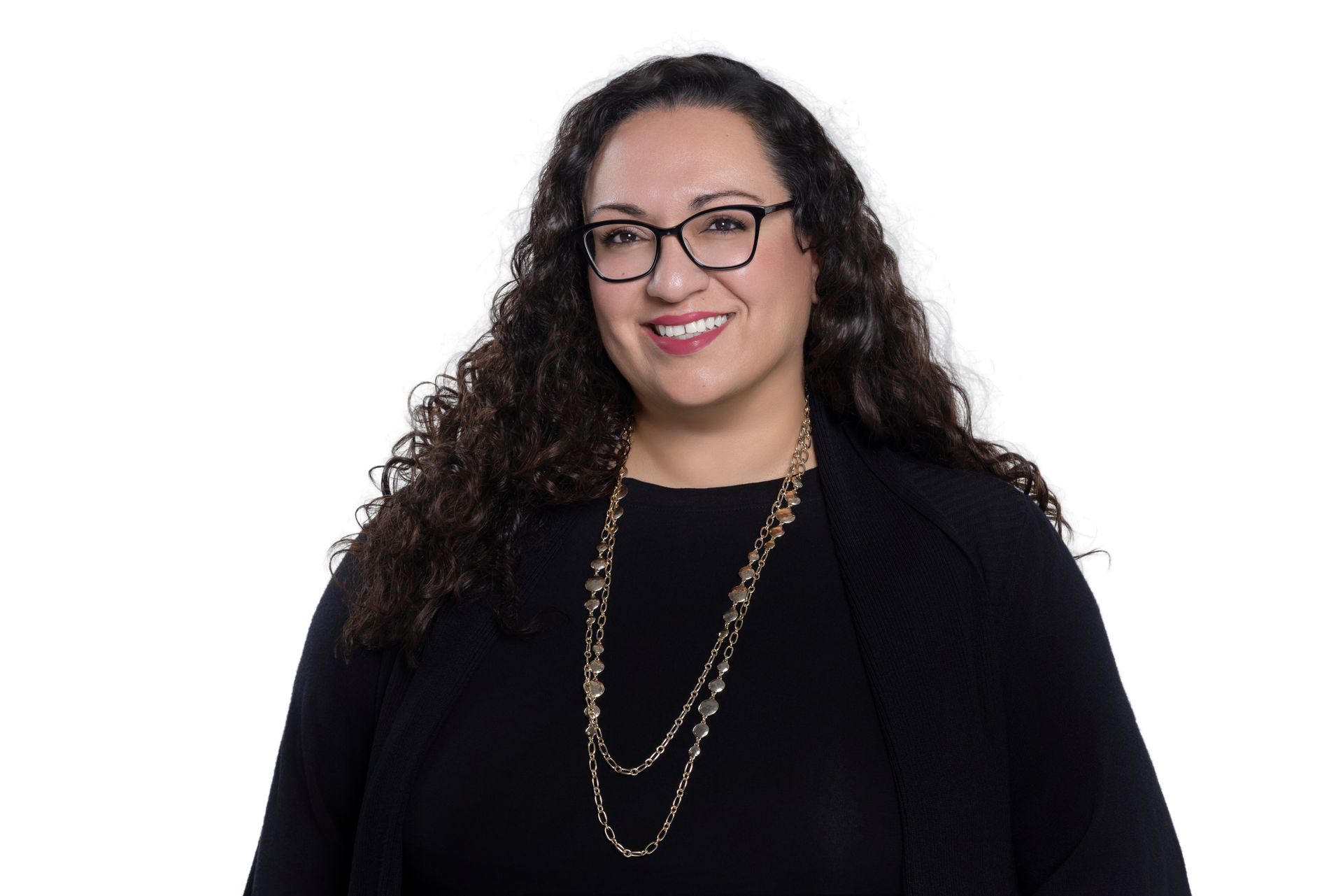 A smiling person with long curly dark hair and glasses, wearing a black top and a long, beaded gold necklace.