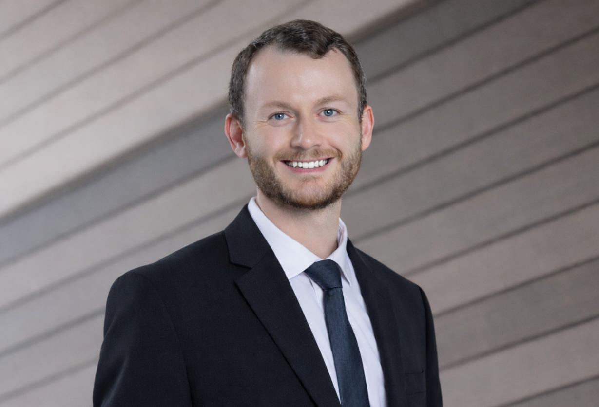 A professional headshot of a smiling person wearing a dark suit, white shirt, and tie against a wood-paneled background.