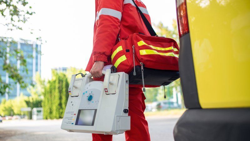 A paramedic is carrying a bag and a device in front of an ambulance.