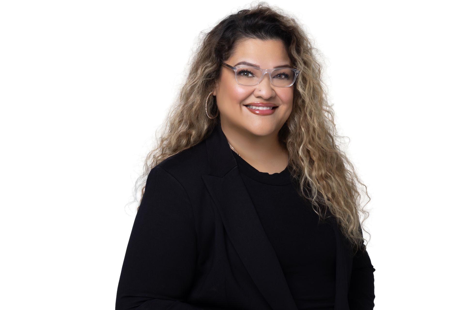 A smiling woman with long wavy hair wearing glasses and a black blazer, isolated against a white background.
