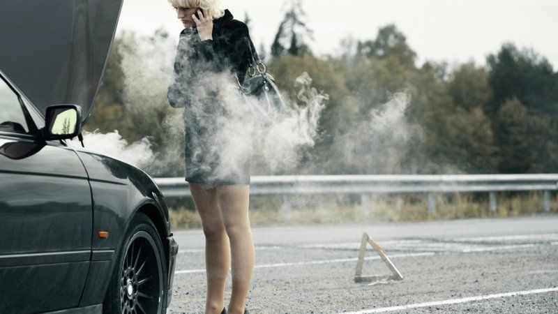 A person stands on the shoulder of a road by a smoking car, speaking on a cell phone with a hazard warning sign nearby.