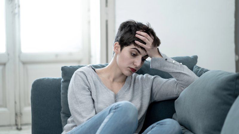 A person with short dark hair sits on a couch, resting their head in one hand with a somber expression.