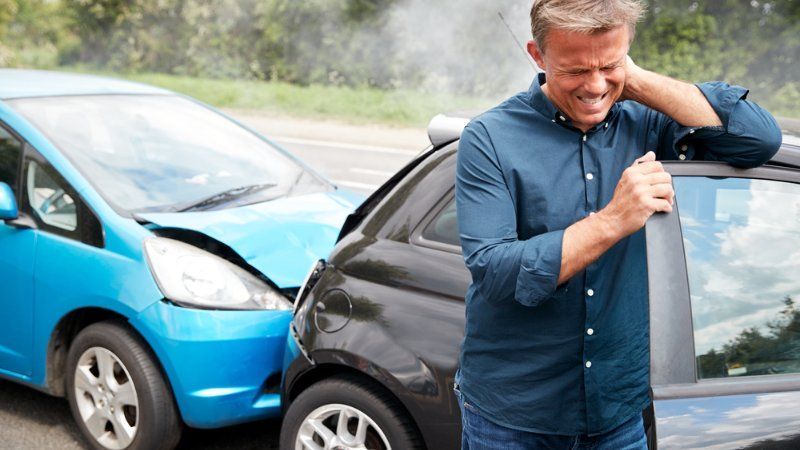 A man in a blue shirt holds his neck in pain while standing next to two cars damaged in a collision.