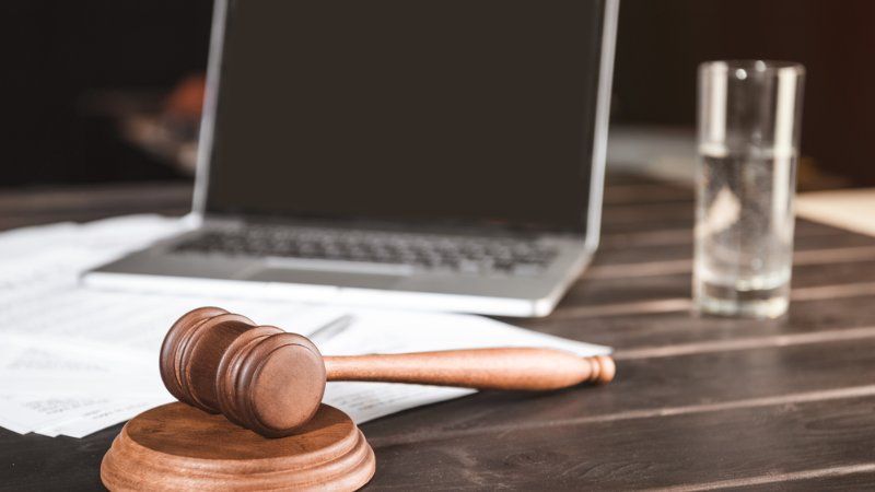 A wooden gavel rests on a round base atop documents, with an open laptop and a glass of water on a desk behind it.