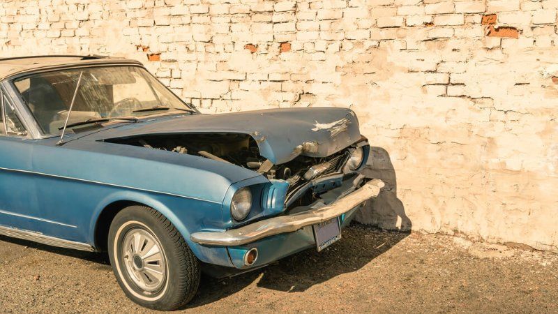 A blue car with its hood open is parked in front of a white brick wall.