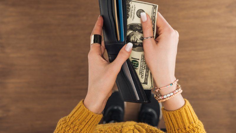 A person wearing a yellow sweater holds an open black wallet with US dollar bills inside against a wooden background.