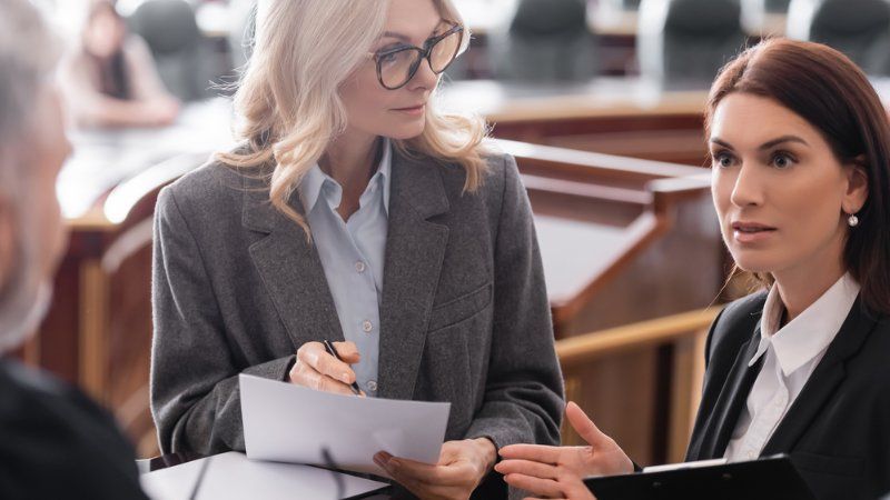 Three professionals in business attire discuss documents in a professional meeting room setting.