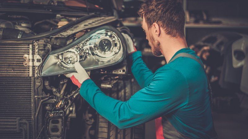 A mechanic in a blue uniform and gloves installing a headlight assembly onto the front of a car in a workshop.