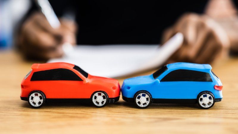 A red and a blue toy car face each other on a desk while a person writes on paper in the background.