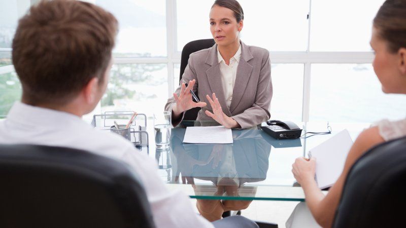 A woman in a blazer gestures while talking to a man and woman seated across from her at a desk in a bright office.