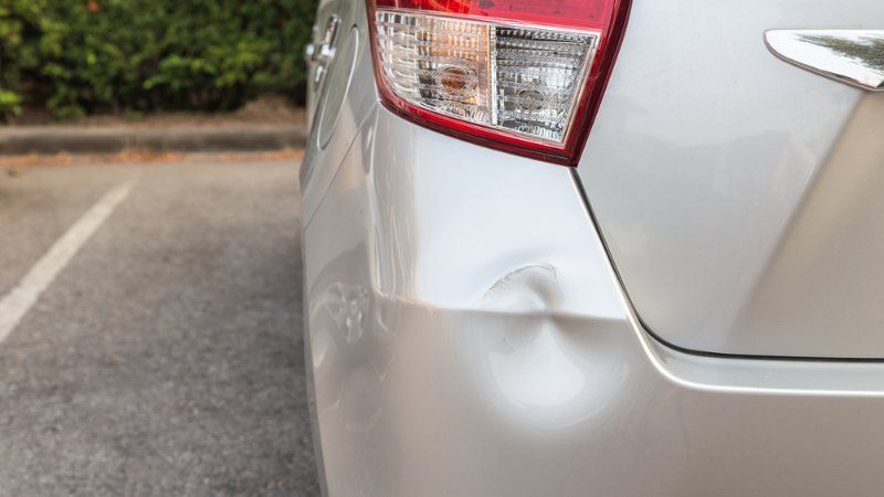A silver car bumper showing a circular indentation or dent below the tail light in a parking lot.