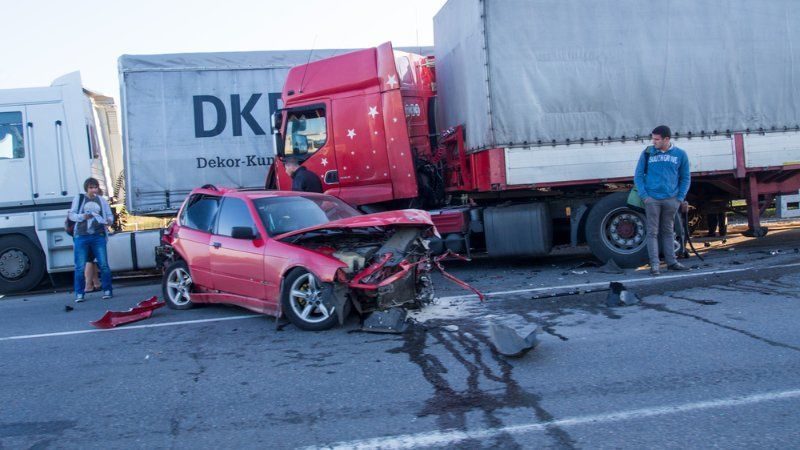 A red car with severe front-end damage sits on a road near two large semi-trucks, with two people standing nearby.