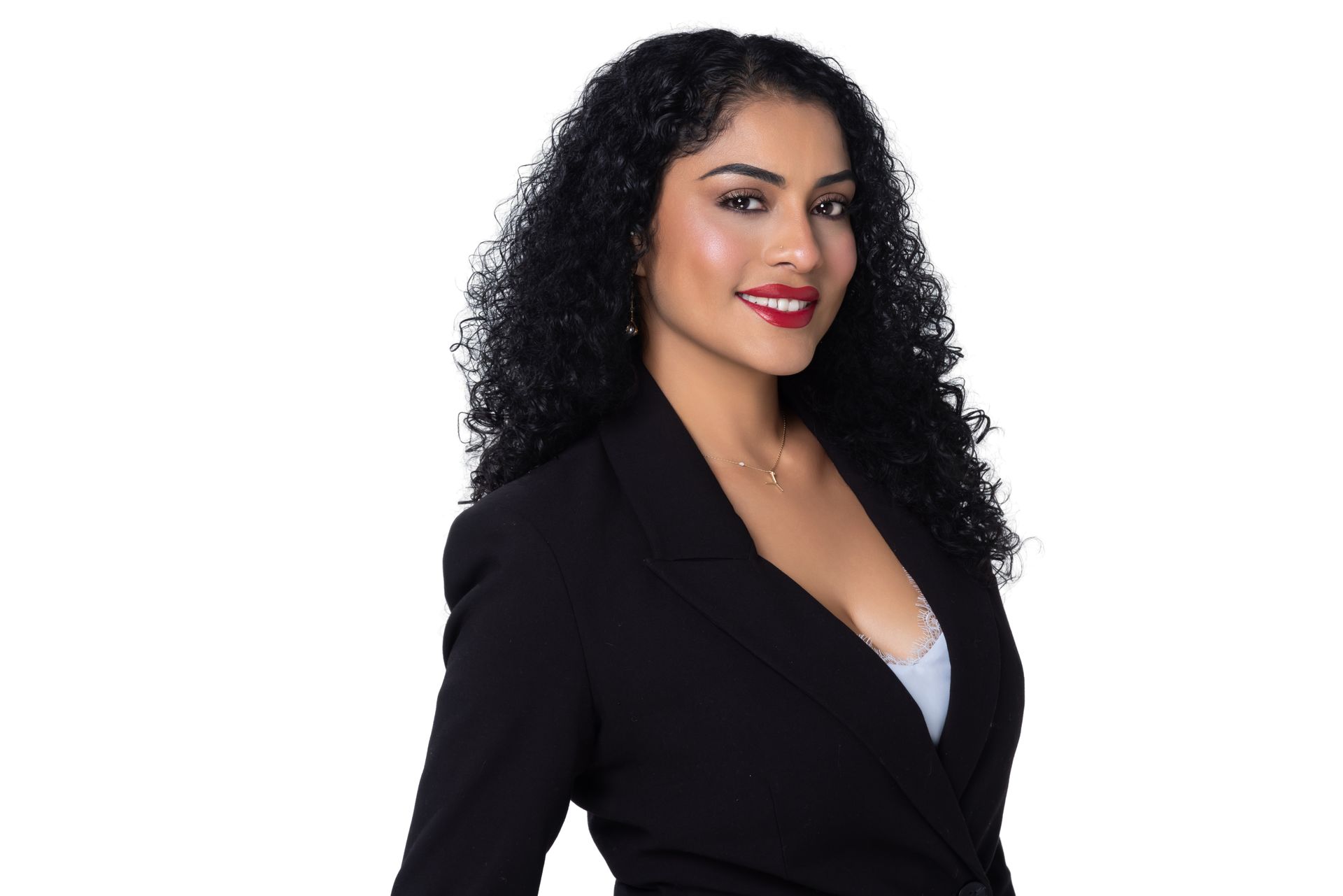 A person with dark curly hair wears a black blazer over a gray top, smiling against a plain white background.