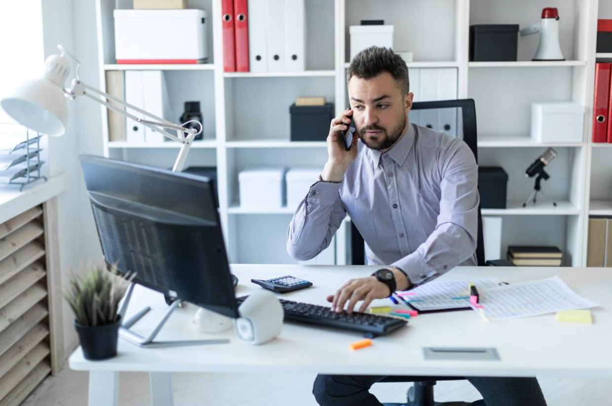 A man is sitting at a desk in front of a computer talking on a cell phone.