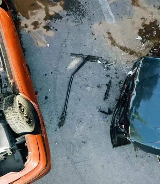 A damaged orange car fender and a dark vehicle bumper lie on wet asphalt, separated by car debris.