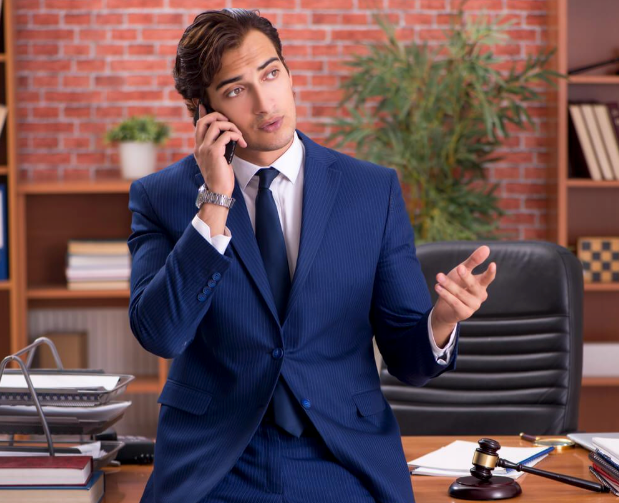 A man in a suit and tie is sitting at a desk talking on a cell phone.