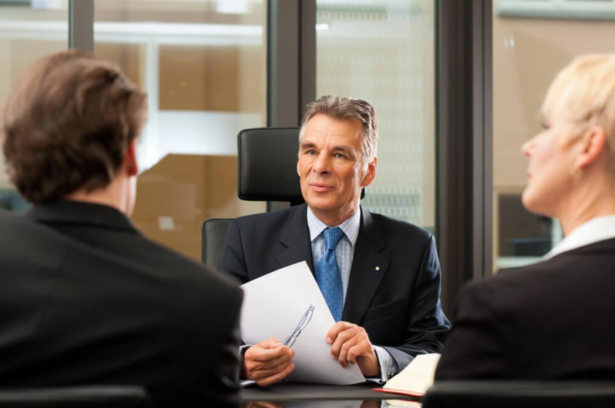 A man in a suit and tie is sitting at a table talking to two people.