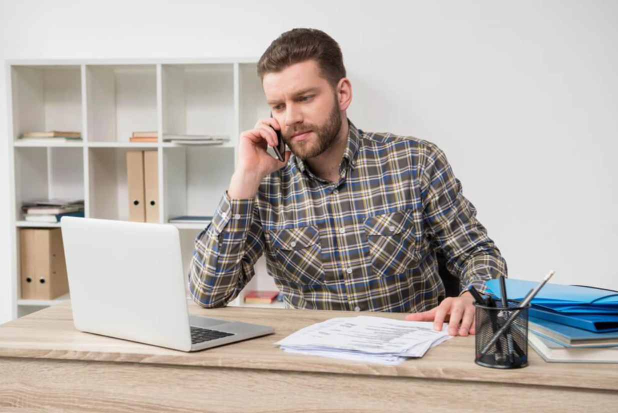 A man is sitting at a desk talking on a cell phone while using a laptop computer.