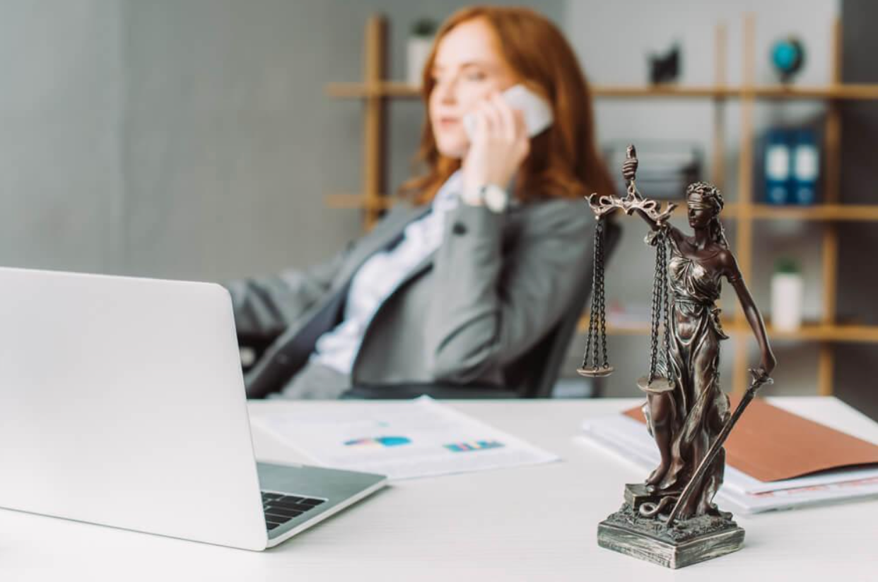 A woman is sitting at a desk talking on a cell phone next to a statue of justice.