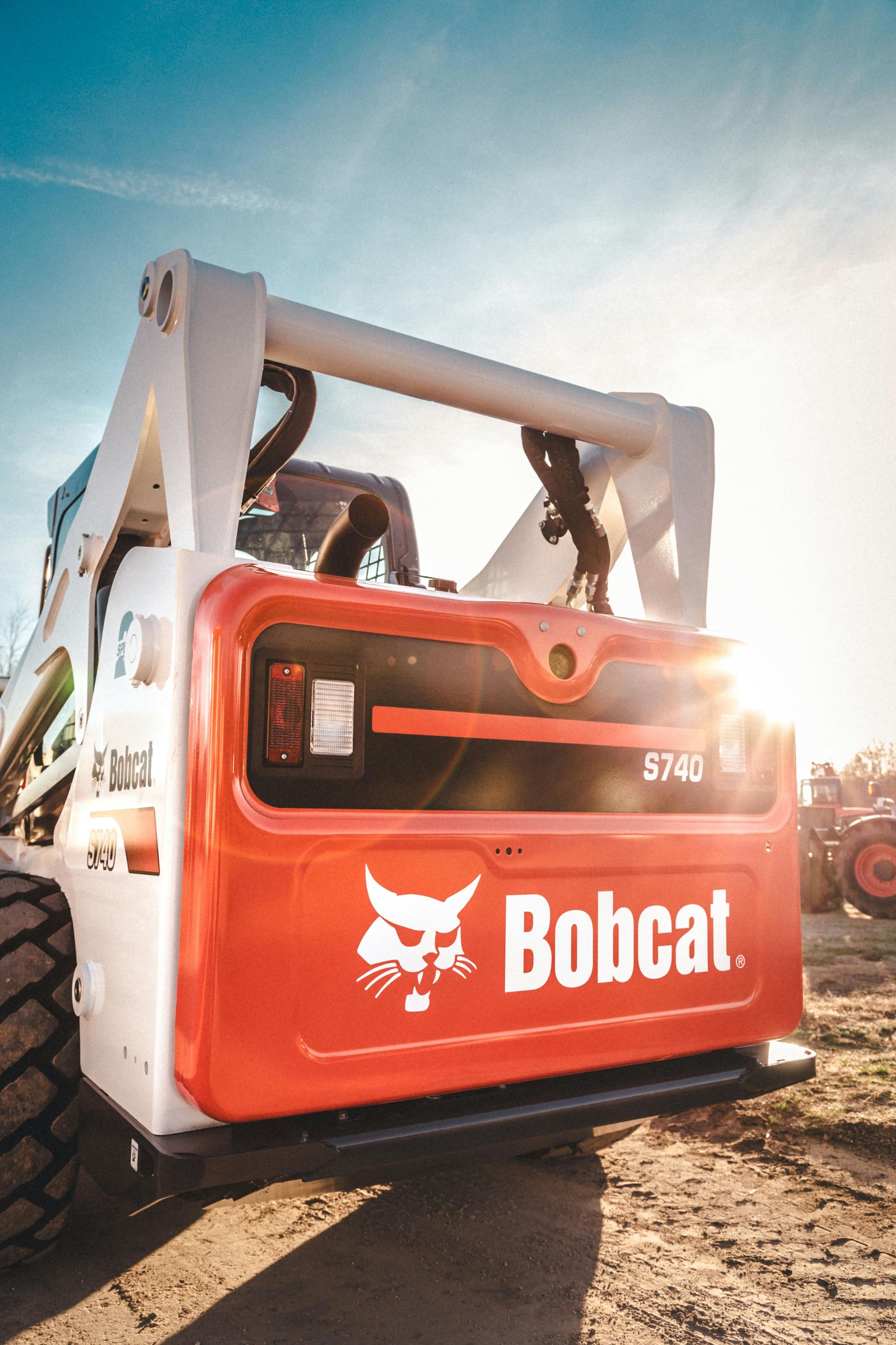 Close-up rear view of an orange and white Bobcat skid-steer loader parked on a dirt lot under a sunny sky.