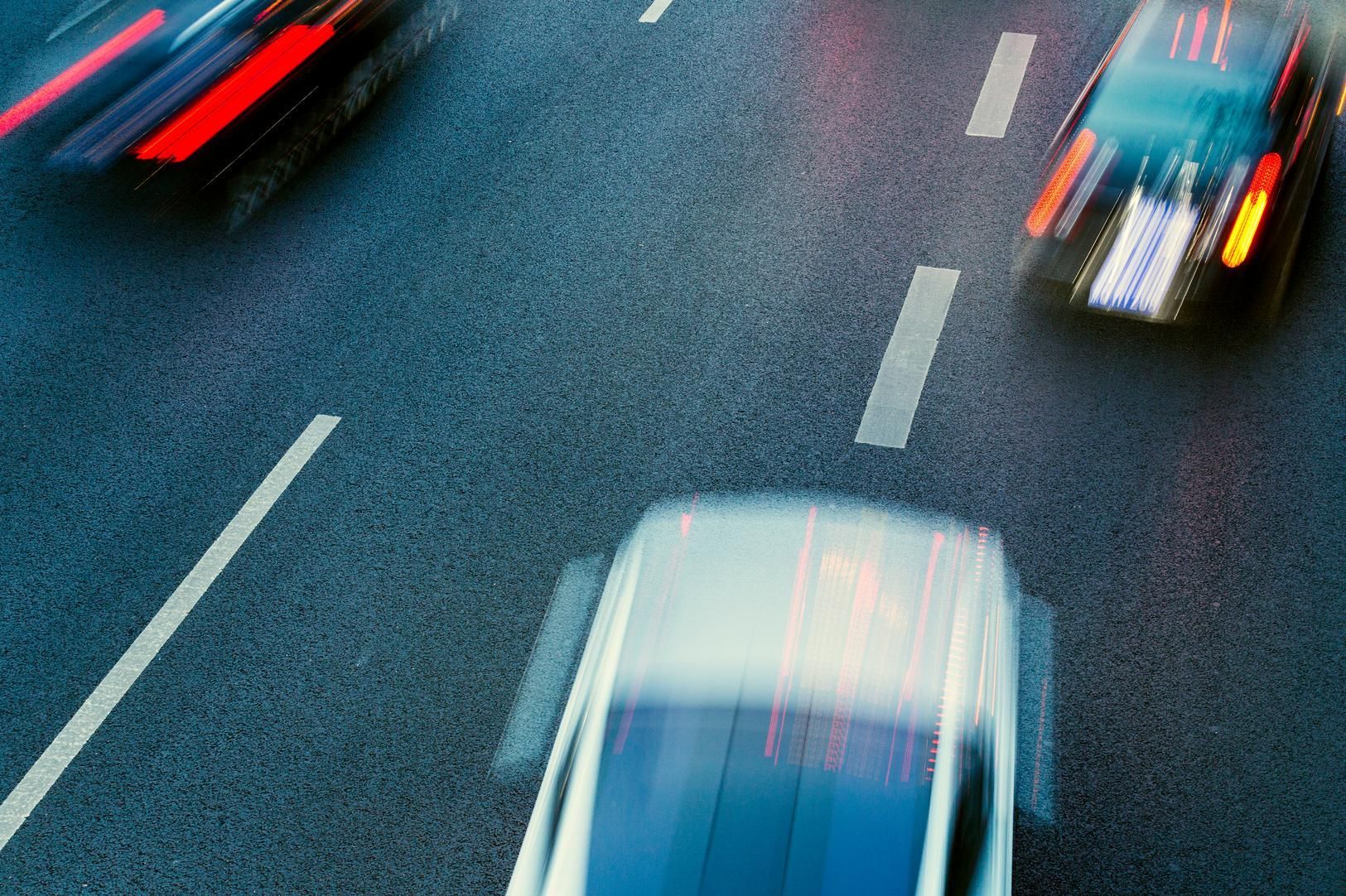 Blurred high-angle view of white and dark cars moving quickly in lanes on a paved highway.